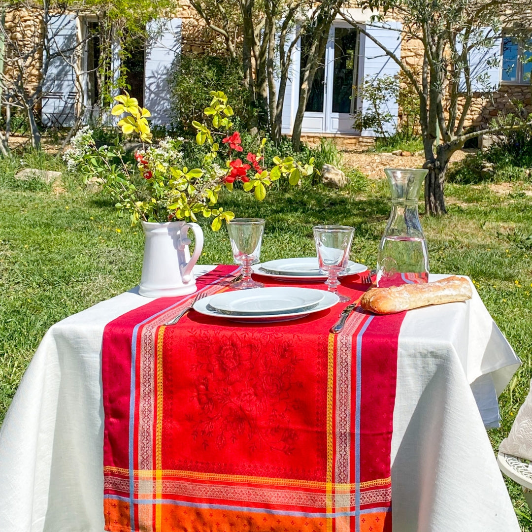 Jacquard‑Tischläufer in intensivem Rot mit beigefarbenen Mustern, gelben Bordüren und hellblauen Akzenten auf einem Esstisch.
